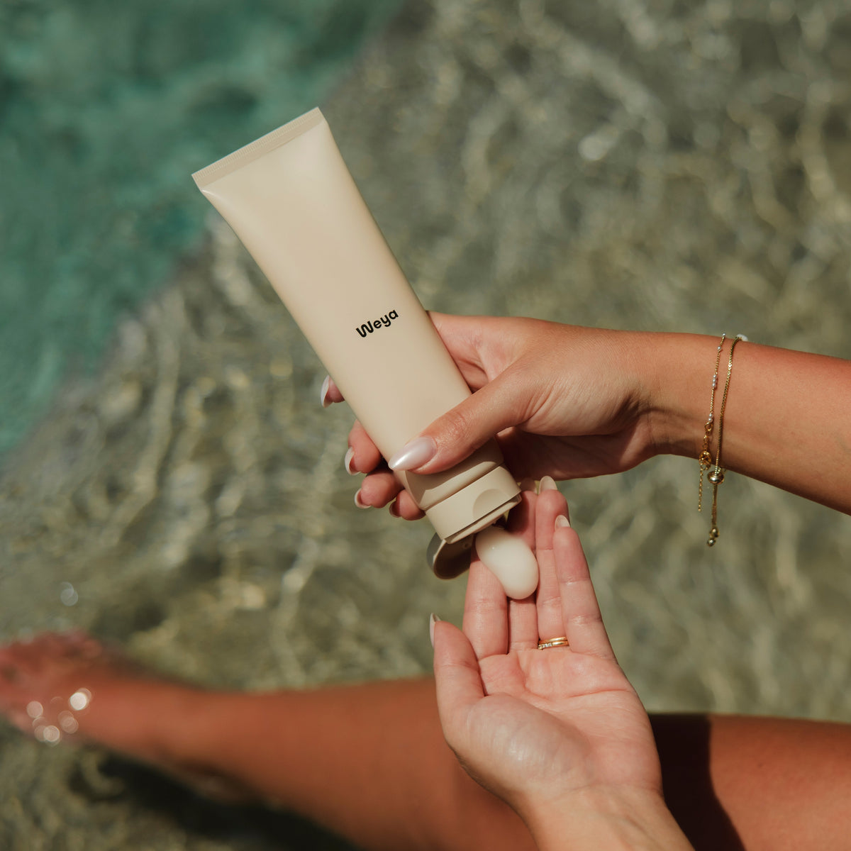 A person's hands and legs are shown holding and dispensing cream from a beige tube labeled 'yepup' in front of a blurred outdoor water background.