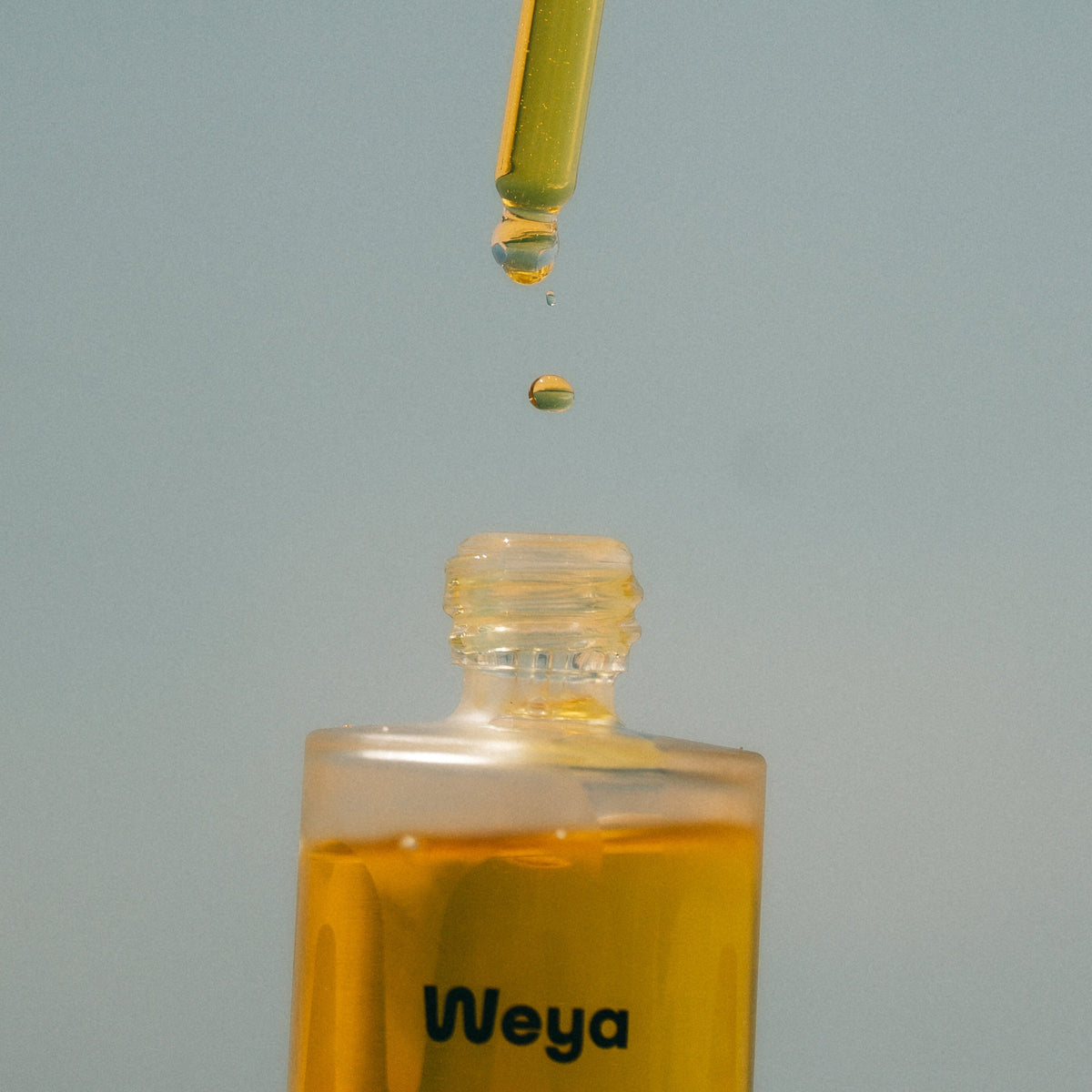 A clear glass dropper bottle with golden-yellow liquid, shown with a dropper above the open bottle and a single drop suspended in mid-air against a soft light blue-gray background. The label reads 'Weya'.