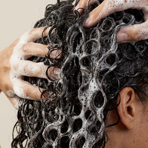 Close-up of curly dark hair being washed with white shampoo lather, with hands visibly soapy and touching the hair. Part of the ear and neck are visible, and the background is a plain warm light beige.