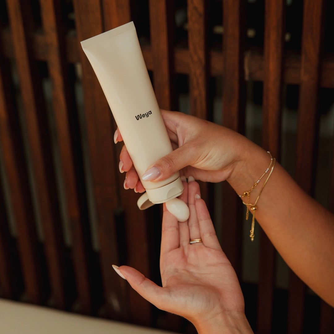 A person's hands holding a beige squeeze tube and dispensing cream onto the other hand, with a dark brown wooden slat background.