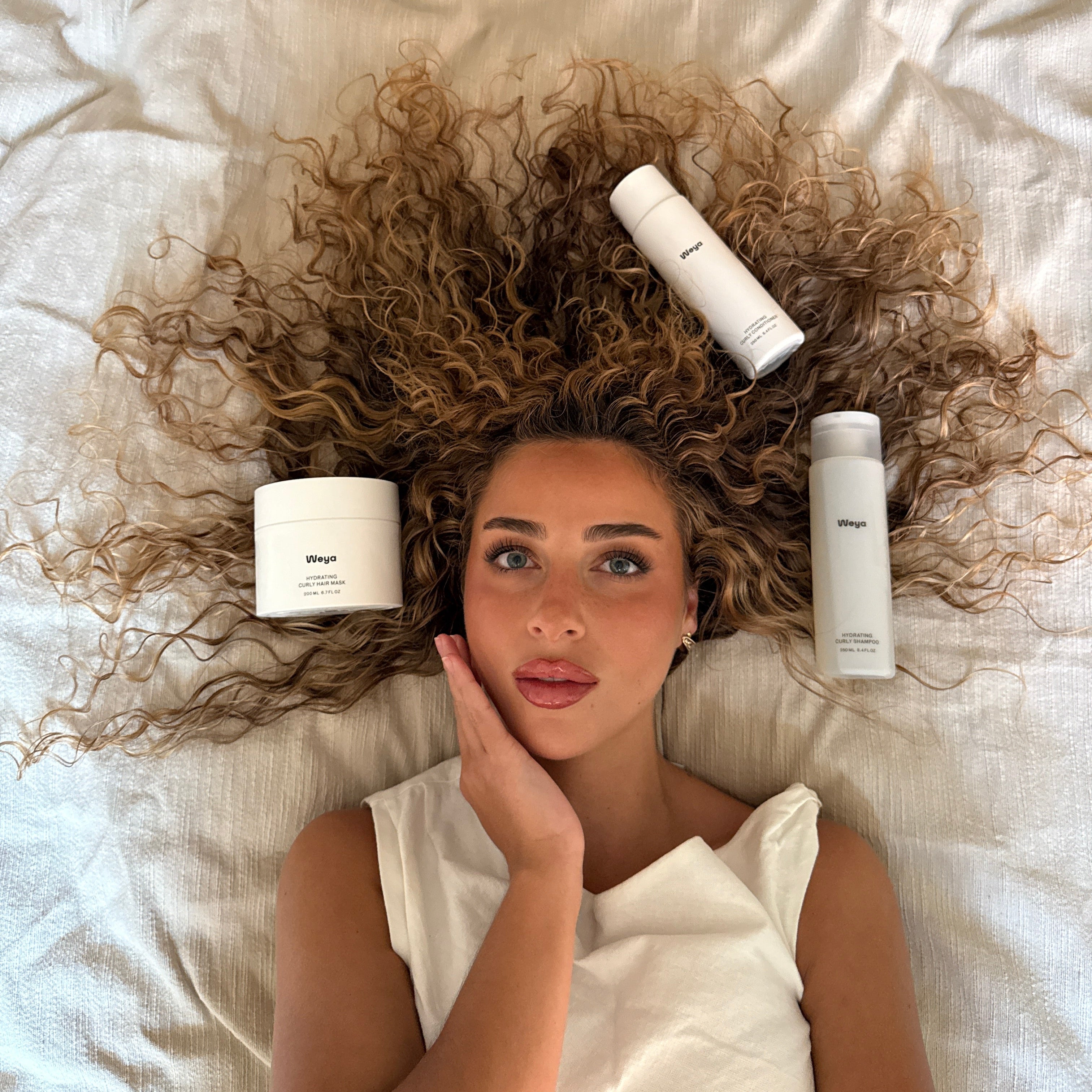 A woman with voluminous curly brown hair lies on white bedding with three white haircare products arranged around her head. The products include a jar, a spray bottle, and a tall bottle, all branded with the word 'Melt'.