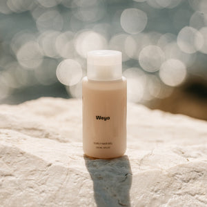 A small beige bottle labeled 'Wega' with a white cap, standing on a light stone surface with a blurred bokeh background.