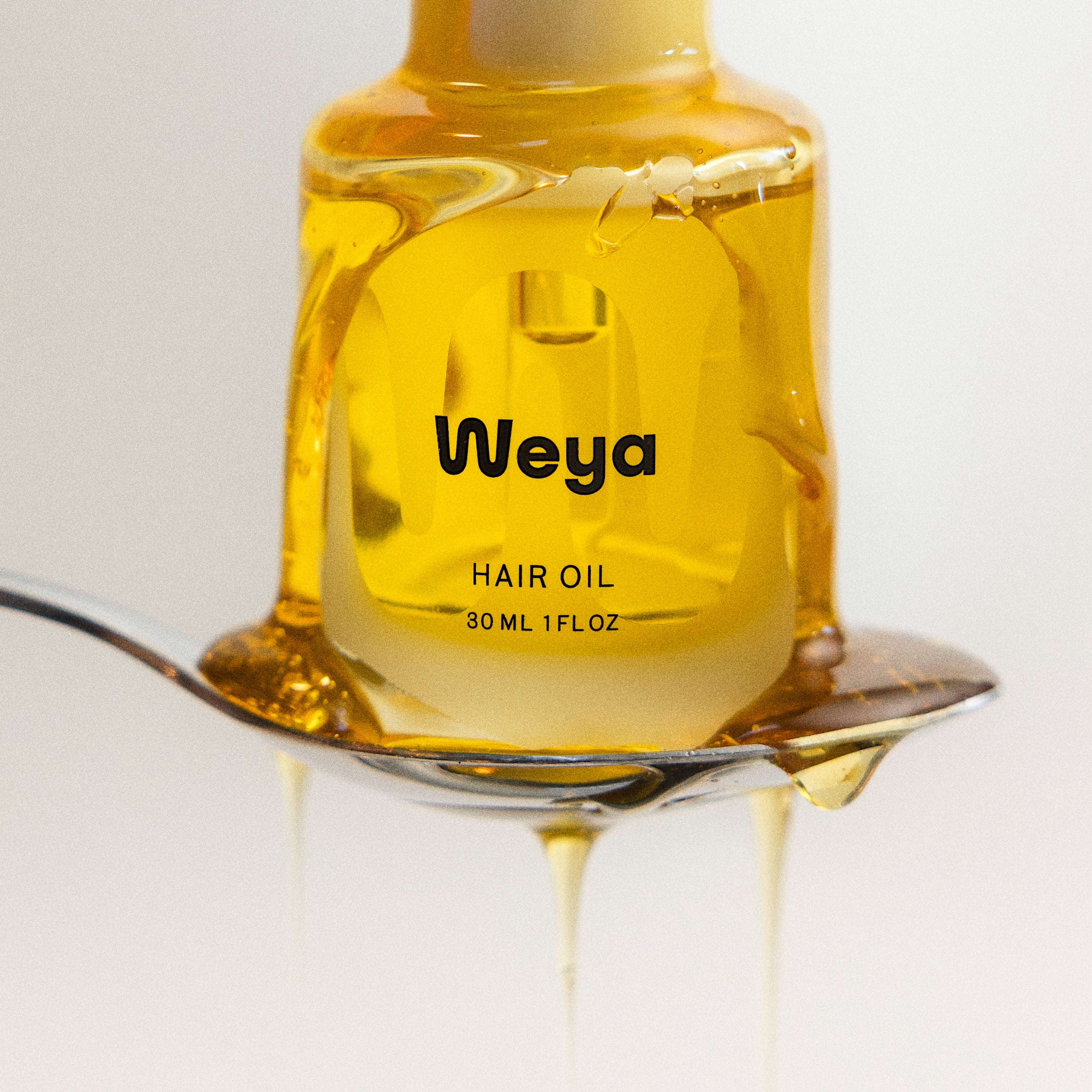 A bottle of Weya hair oil filled with golden yellow oil, held on a metal spoon with oil dripping down, against a clean white background.