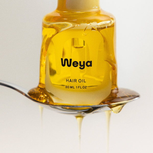 A bottle of Weya hair oil filled with golden yellow oil, held on a metal spoon with oil dripping down, against a clean white background.