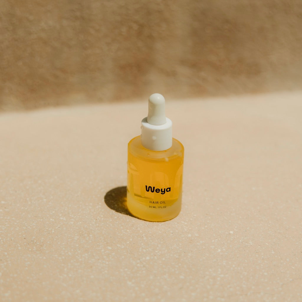 A small dropper bottle of Weya facial oil filled with golden-yellow liquid, standing on a sandy beige surface with a soft-focus textured background.