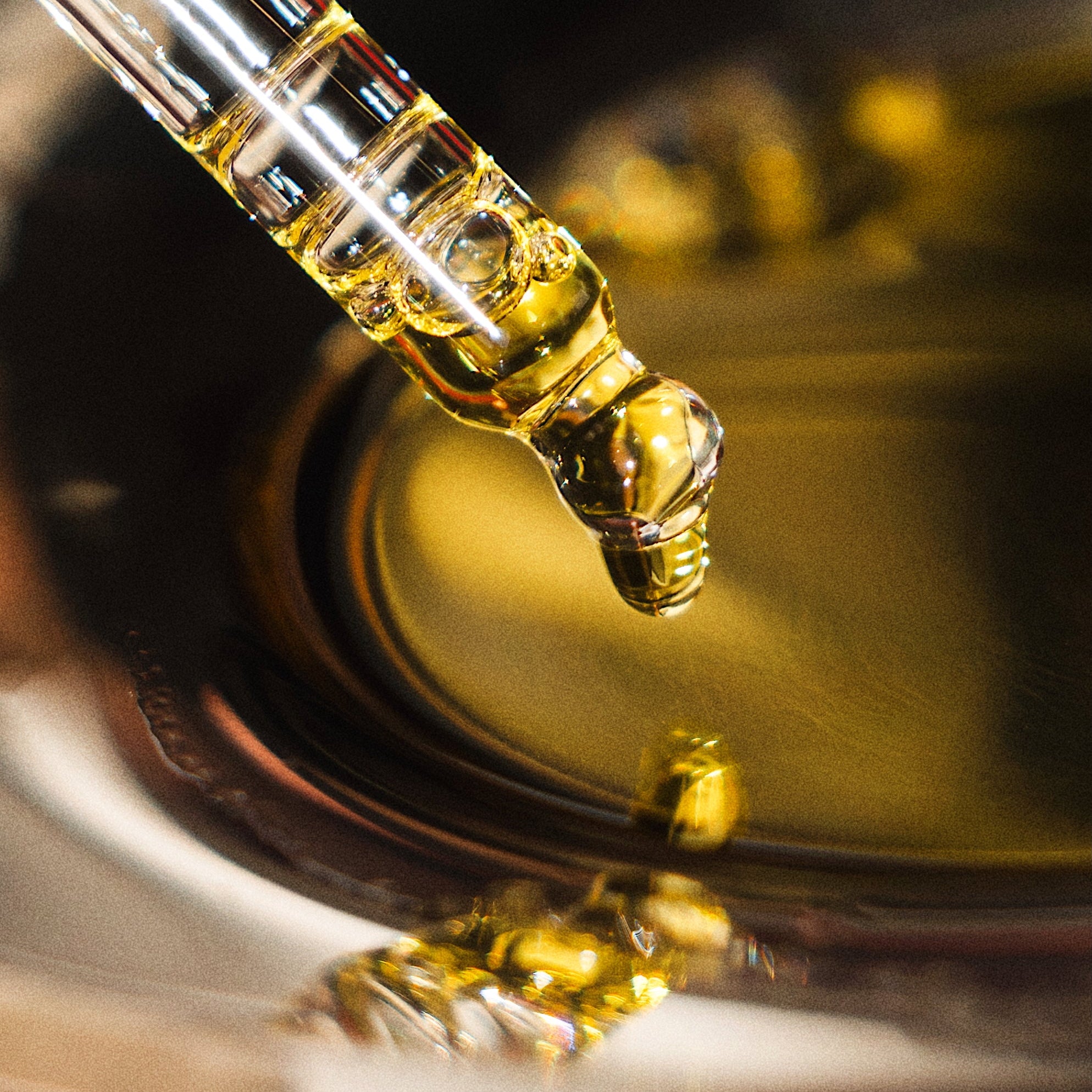A close-up of a glass dropper containing golden yellow oil, positioned above a reflective metal container filled with the same oil.