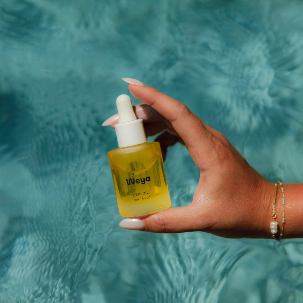 A hand holding a small bottle of Weya hair oil with a dropper cap, set against a blurred background of rippling blue water.