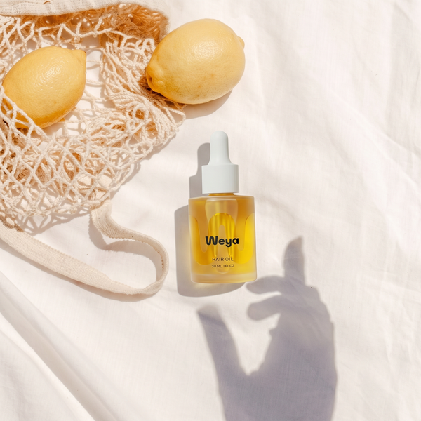 Bottle of Weya hair oil with lemons and a mesh bag on a white background