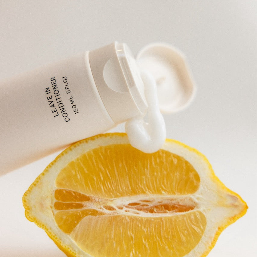 A white squeeze tube of hair conditioner with white cream dripping out, positioned above a halved lemon on a clean white background. The tube has black text reading 'LEAVE-IN HAIR CONDITIONER'.