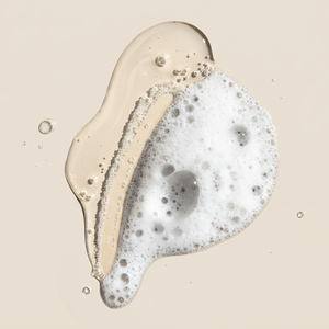 A close-up studio shot of a translucent beige liquid with white foam and small bubbles on a warm beige background.